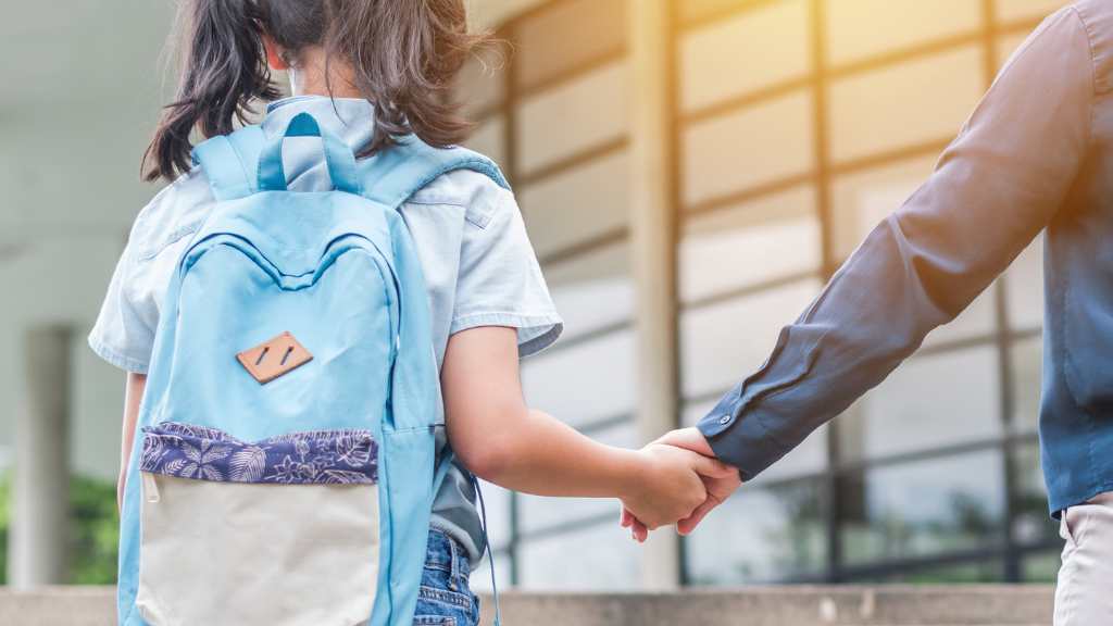 Niña con mochila azul entrando al colegio mientras sostiene la mano de un adulto. Imagen representativa del regreso a clases y el apoyo familiar en el manejo de la ansiedad en niños con autismo. Ideal para ilustrar temas como autismo y ansiedad escolar, adaptación escolar y estrategias para reducir la ansiedad en niños con TEA.